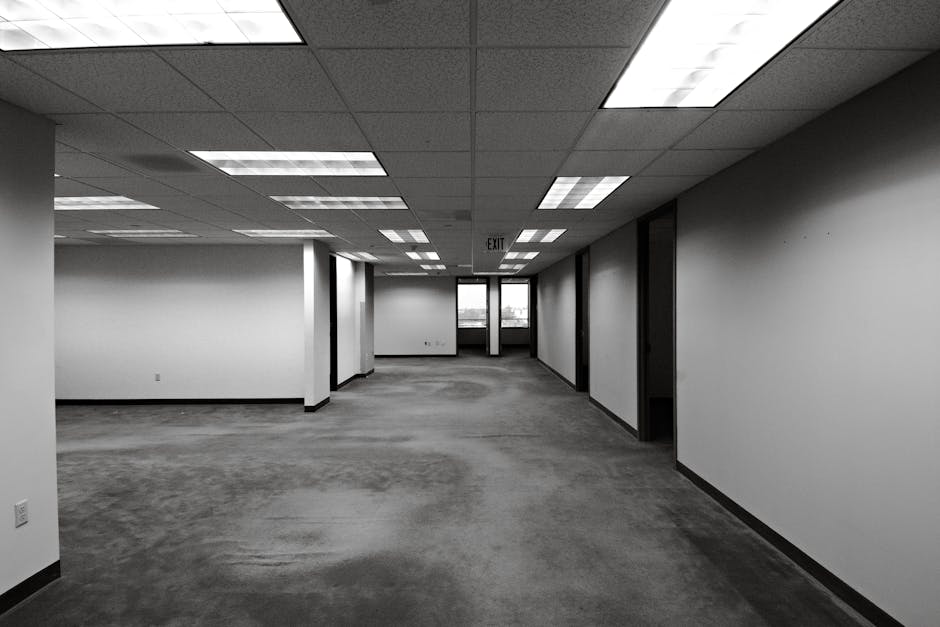 Black and white image of an empty office space with fluorescent lighting and exit sign.